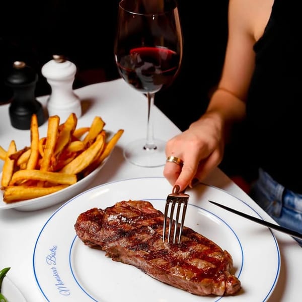 A hand cuts into a grilled steak on a Monsieur Bistro plate with fries and red wine.