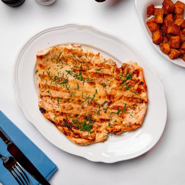 Grilled chicken breast with herbs on a white plate, served with roasted potatoes and cutlery.