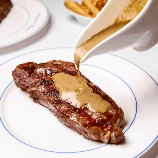 Peppercorn sauce being poured from a white gravy boat onto a grilled steak on a white plate.